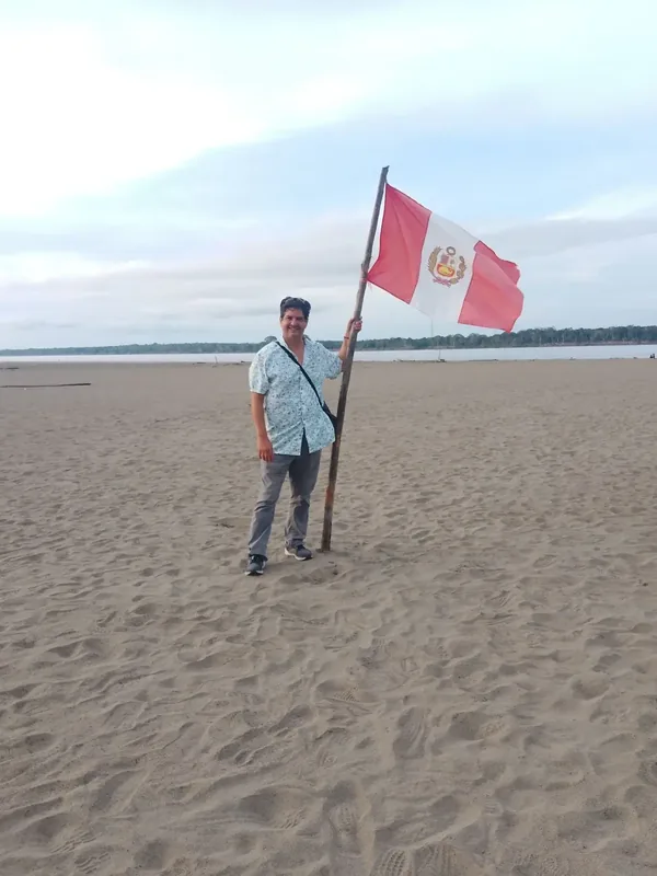 Miguel standing beside a riverside beach in Peru with the Peruvian flag