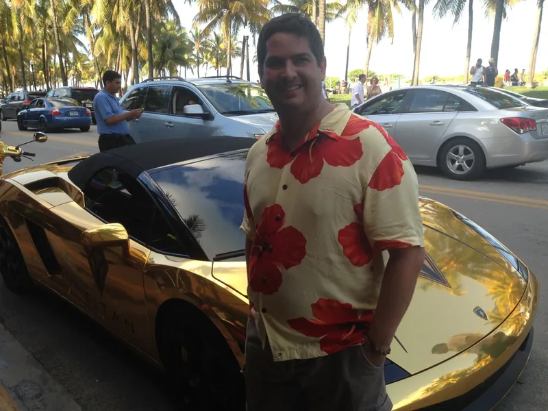 Miguel beside a gold car in a sunny Caribbean street scene