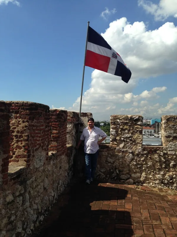 Miguel standing beside a Dominican flag in a historic colonial fort